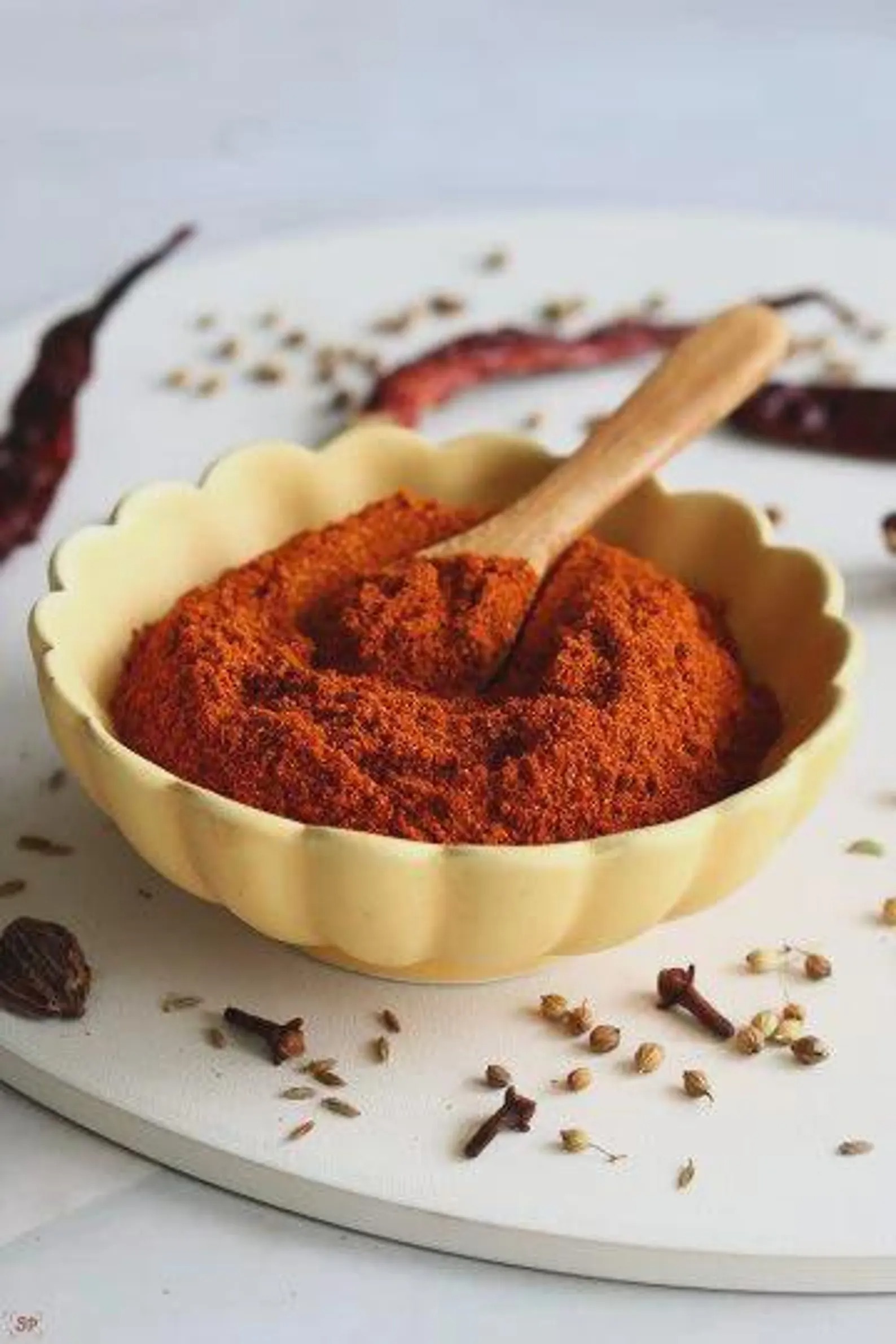 Bowl of deep red chilli and spice blend with a wooden spoon, surrounded by whole spices